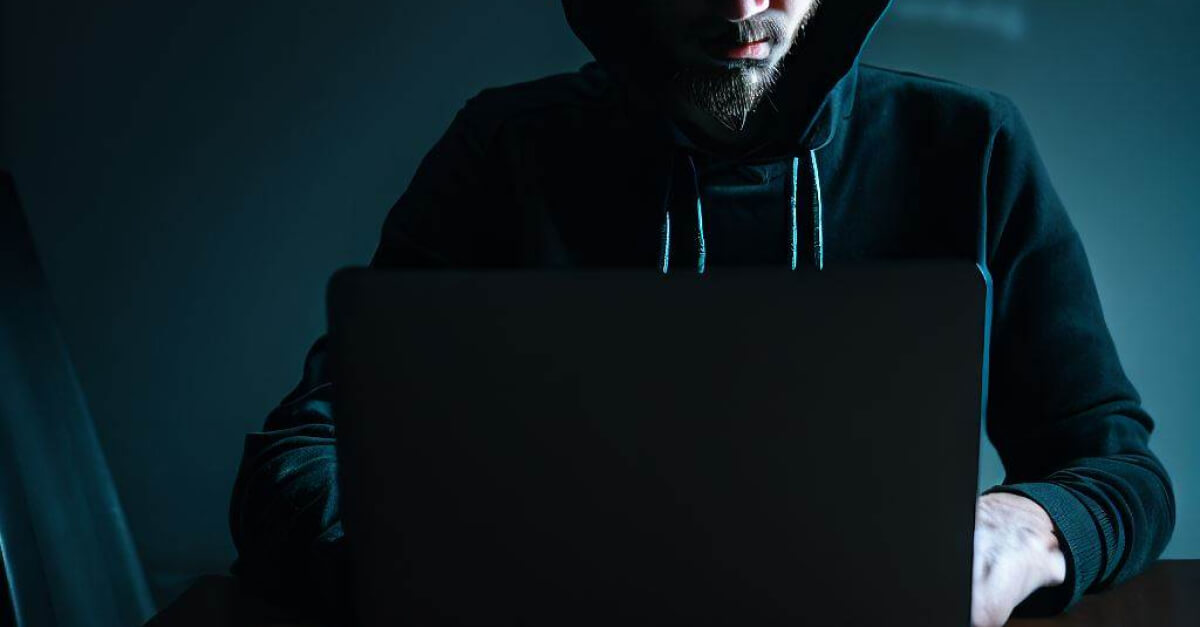 Seorang pria sedang di depan laptop bergaya seperti seorang hacker di ruangan yang gelap